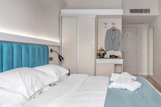 A double bed with 2 neatly folded bathrobes, wardrobes, mini bar and fresh flowers in a vase on top. A view of entry/exit door of the room.