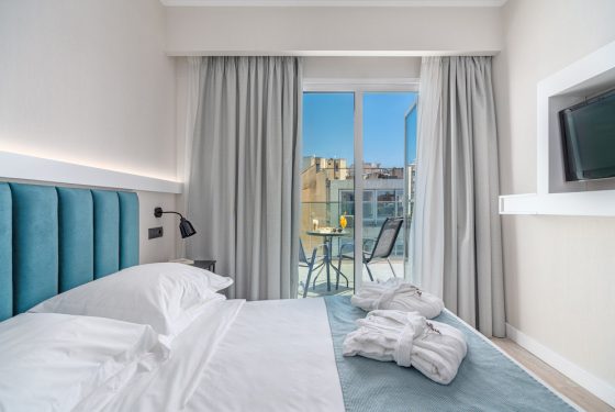 Double bed with 2 neatley folded bathrobes. A view of the city from open draped balcony doors. A table with 2 chairs placed on the balcony.