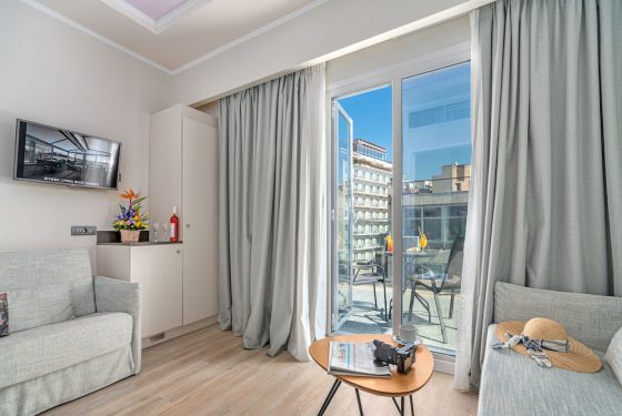 2 sofas facing each other with a small table in the center. On top of the table is a coffee cup, newspaper & camera. A tv hangs on the wall next to the mini bar. A bottle of wine, 2 glasses & fresh flowers are placed on top. Open draped balcony doors overlooking a view of the city.